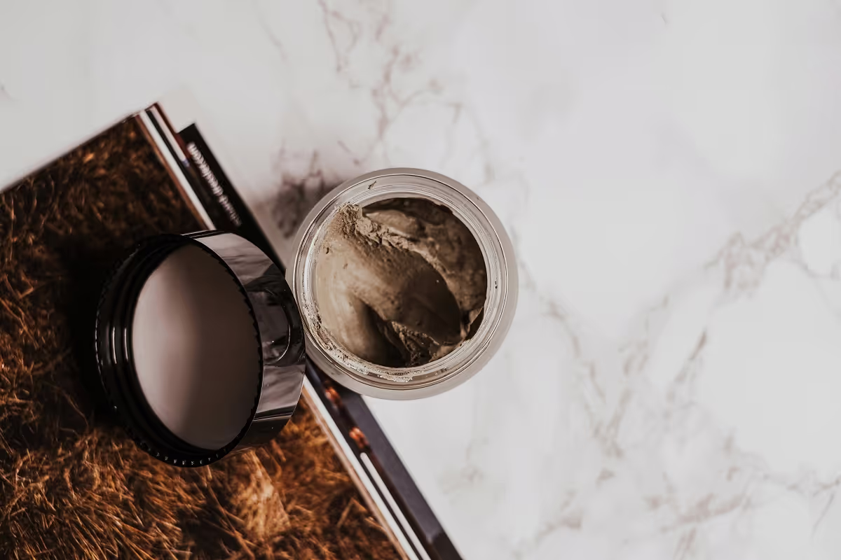 An open jar of clay mask beside its black lid and a magazine on a marble tabletop